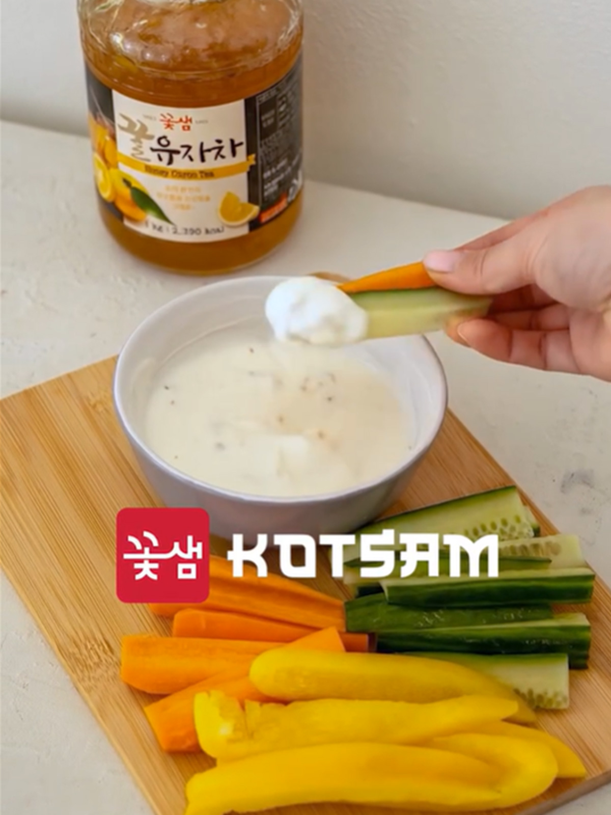 Letní citrusový dip s Kotsam yuzu v misce na dřevěném prkénku; mrkvové a okurkové tyčinky, plátky papriky; v pozadí rozostřený produkt Kotsam, logo uprostřed