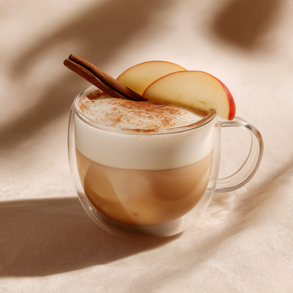 Dvoustěnný skleněný šálek s mléčným nápojem z Kotsam Apple Tea; krémová barva s jemnými čajovými víry, mikro pěna, plátek jablka a skořicová tyčinka