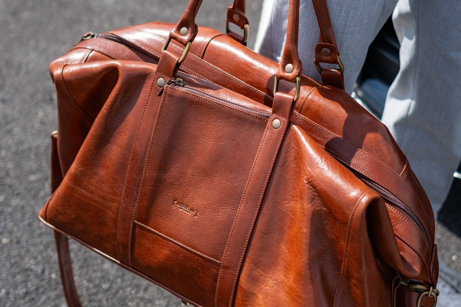 Detail lesklé koňakové usně tašky Marco, kožený pás s nýty a zip se staromosazným kováním