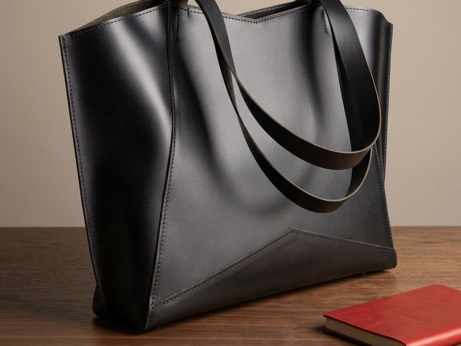 Černá kožená taška Margot Shopper ze tříčtvrtečního pohledu na ořechovém stole, vedle červený kožený zápisník