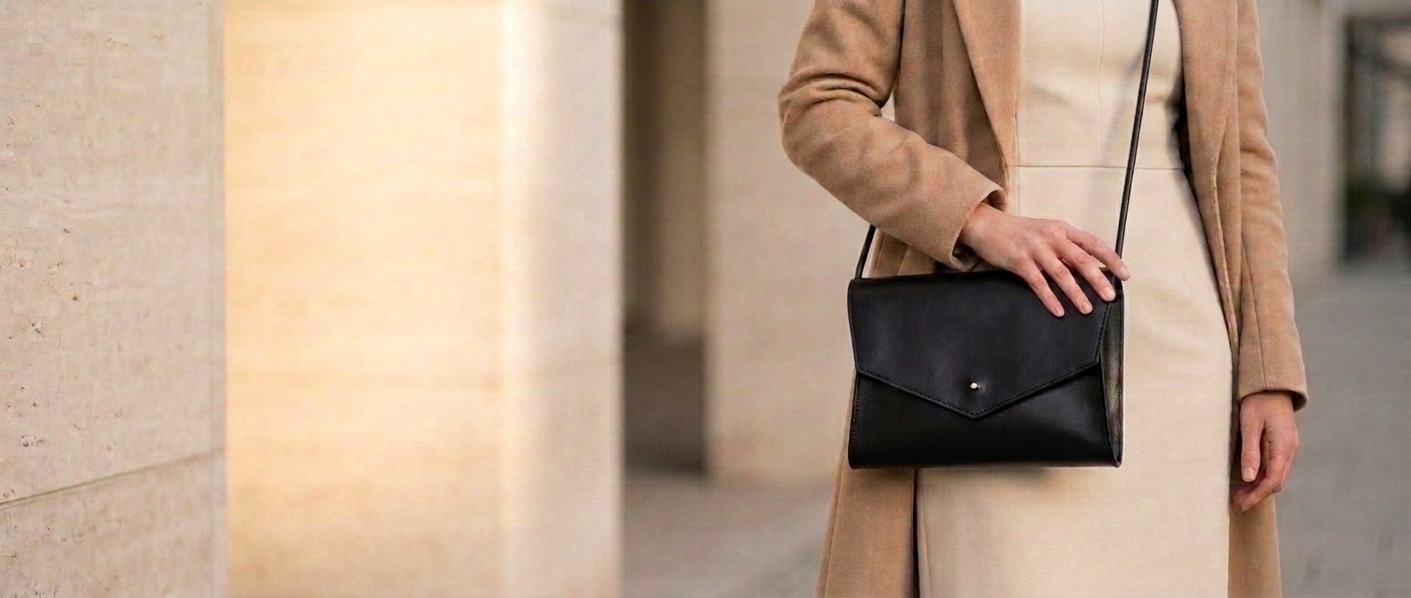 Žena v krémových šatech a kabátě nese černou koženou kabelku Mia 2.0 crossbody na tenkém popruhu