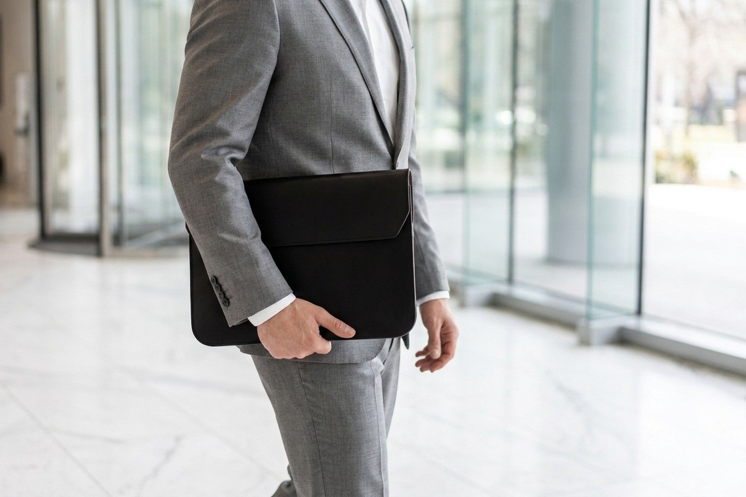 Muž v světle šedém obleku nese černé pouzdro na notebook pod paží moderní halou s mramorovou podlahou