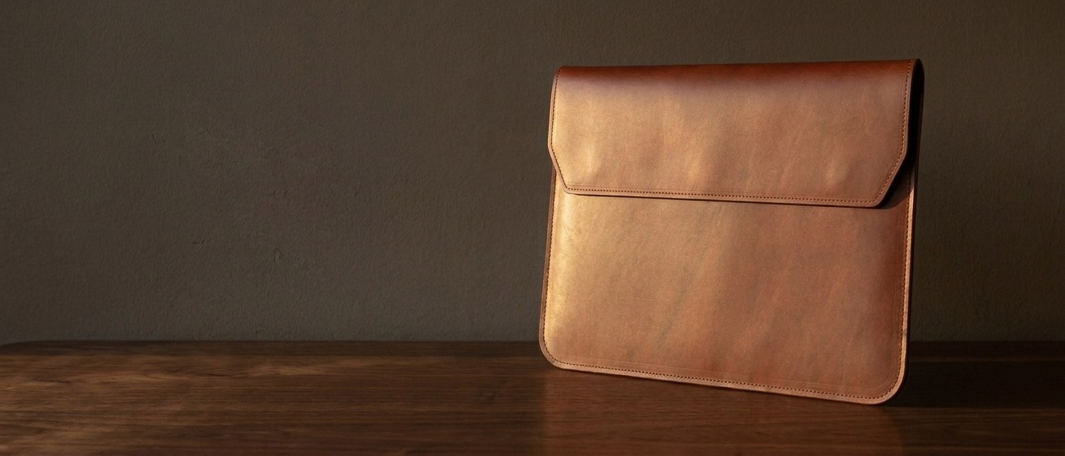 Hnědé kožené pouzdro na notebook 14″ z usně Buttero na tmavém ořechovém stole, vedle hrana notebooku
