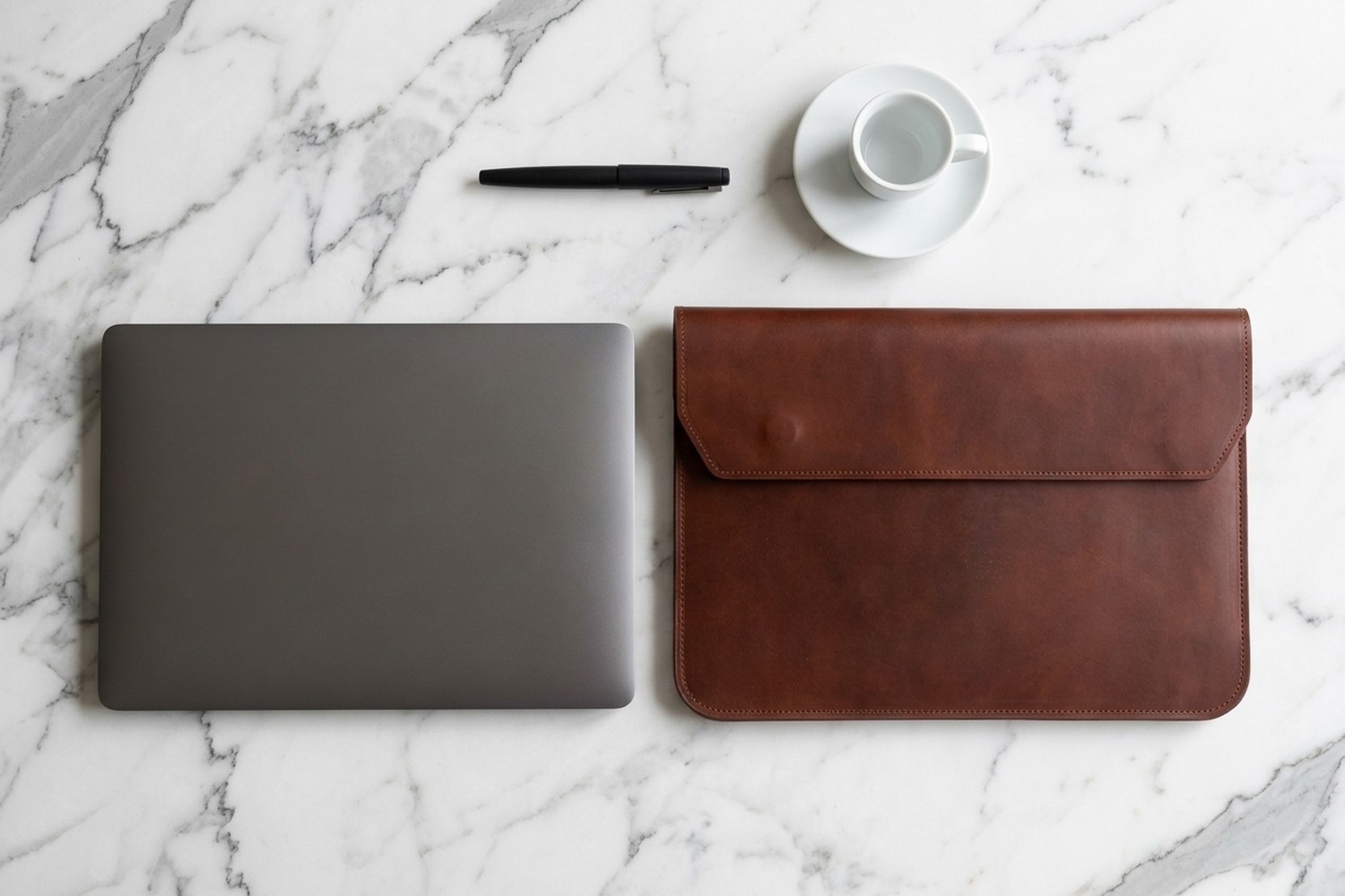 Hnědé pouzdro vedle notebooku 14″ na mramorovém stole, plnicí pero a espresso
