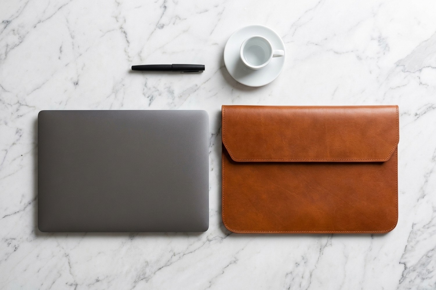 Koňakové pouzdro vedle notebooku 14″ na mramorovém stole, plnicí pero a espresso