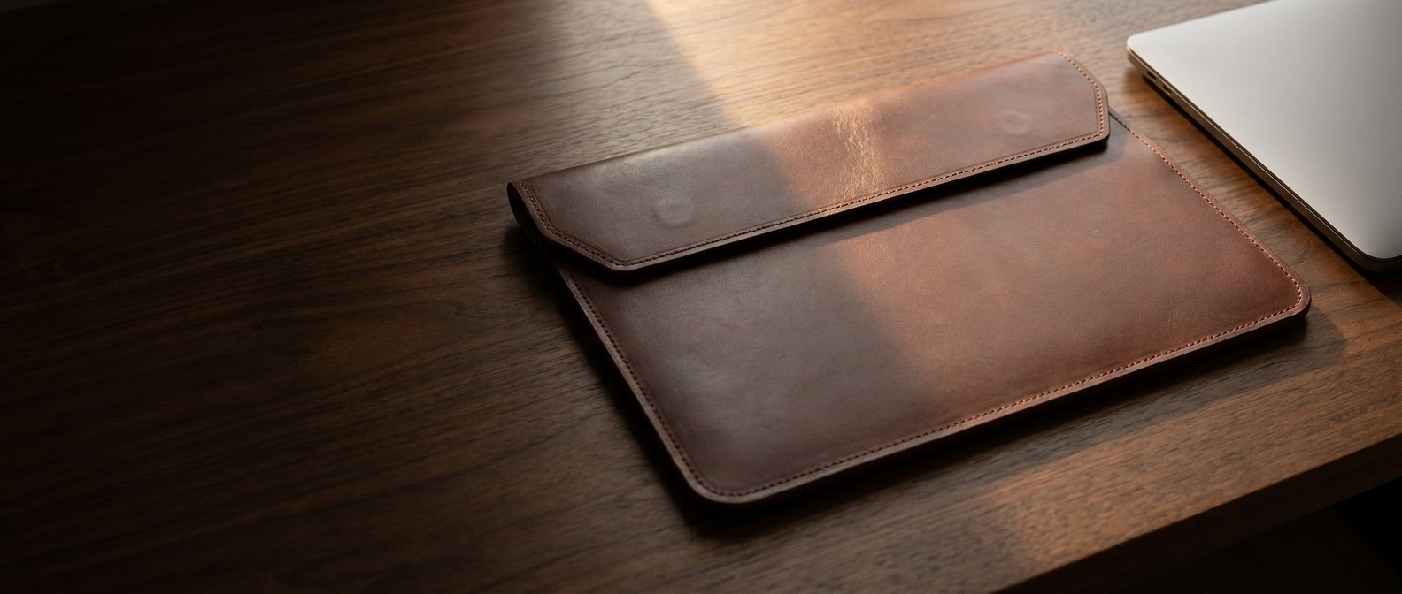 Hnědé kožené pouzdro na tablet z usně Buttero na tmavém ořechovém stole, vedle hrana notebooku