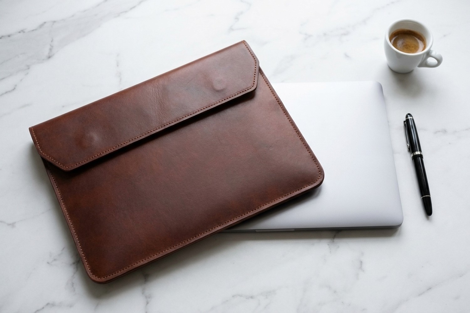 Hnědé pouzdro vedle zavřeného notebooku na mramorovém stole, plnicí pero a espresso