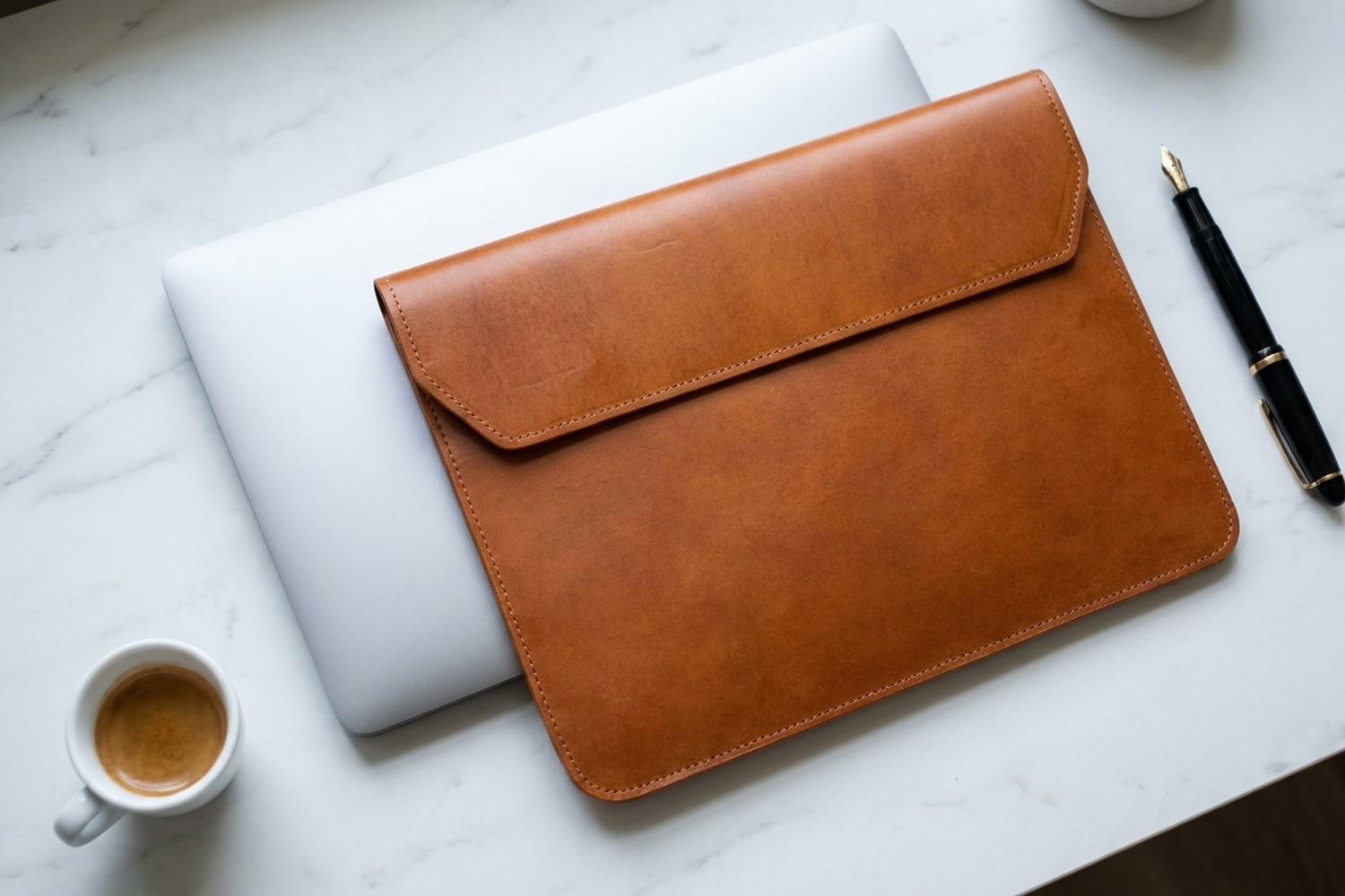 Koňakové pouzdro vedle zavřeného notebooku na mramorovém stole, plnicí pero a espresso
