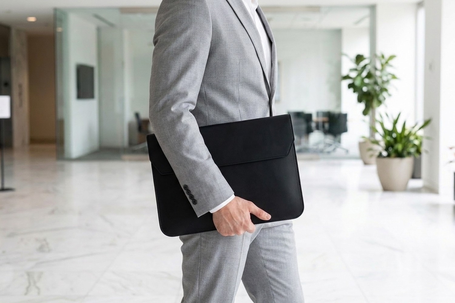 Muž v světle šedém obleku nese černé pouzdro na notebook pod paží moderní halou s mramorovou podlahou