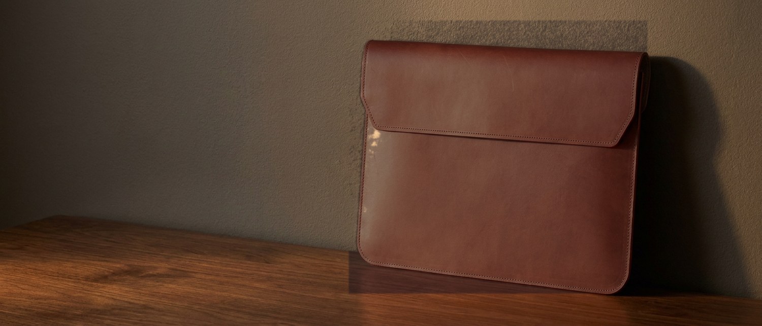 Hnědé kožené pouzdro na notebook 15″ z usně Buttero na tmavém ořechovém stole, vedle hrana notebooku
