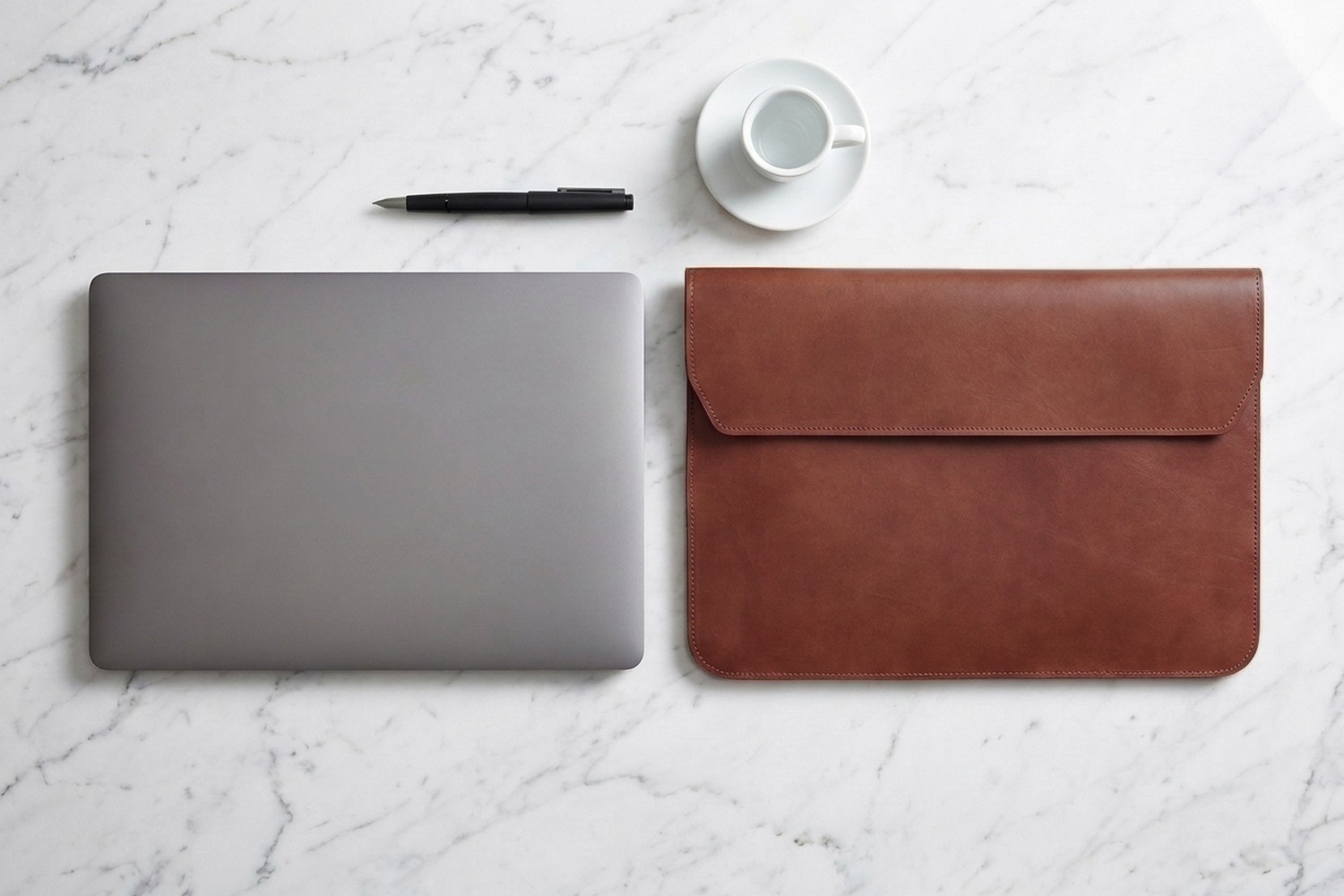 Hnědé pouzdro vedle notebooku 15″ na mramorovém stole, plnicí pero a espresso