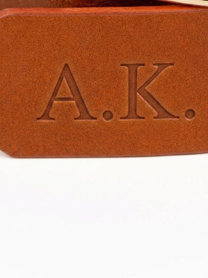 Monogram vyražený do špice koňakového opasku, slepá ražba bez barvy