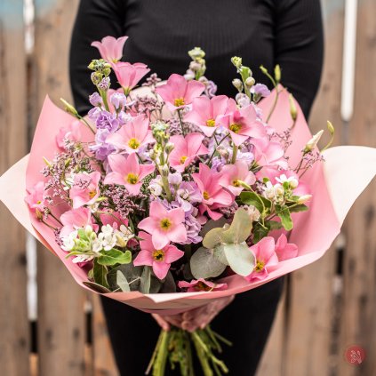 Ručně svázaná kytice obsahující růžovou eustomu, fialovou mathiolu, limonium, oxypetalum a eukalyptové listy, obklopená růžovým obalem, držená ženou v černém svetru