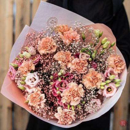 Ručně svázaná kytice s růžovými karafiáty, eustoma a jemným nevěstiným závojem, obklopená jemně růžovým obalem, držená ženou v černém kabátu