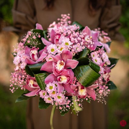 Luxusní růžová kytice s orchidejemi cymbidium, chryzantemami santini, aspidistrou a gypsophilou, držená ženou v zahradě