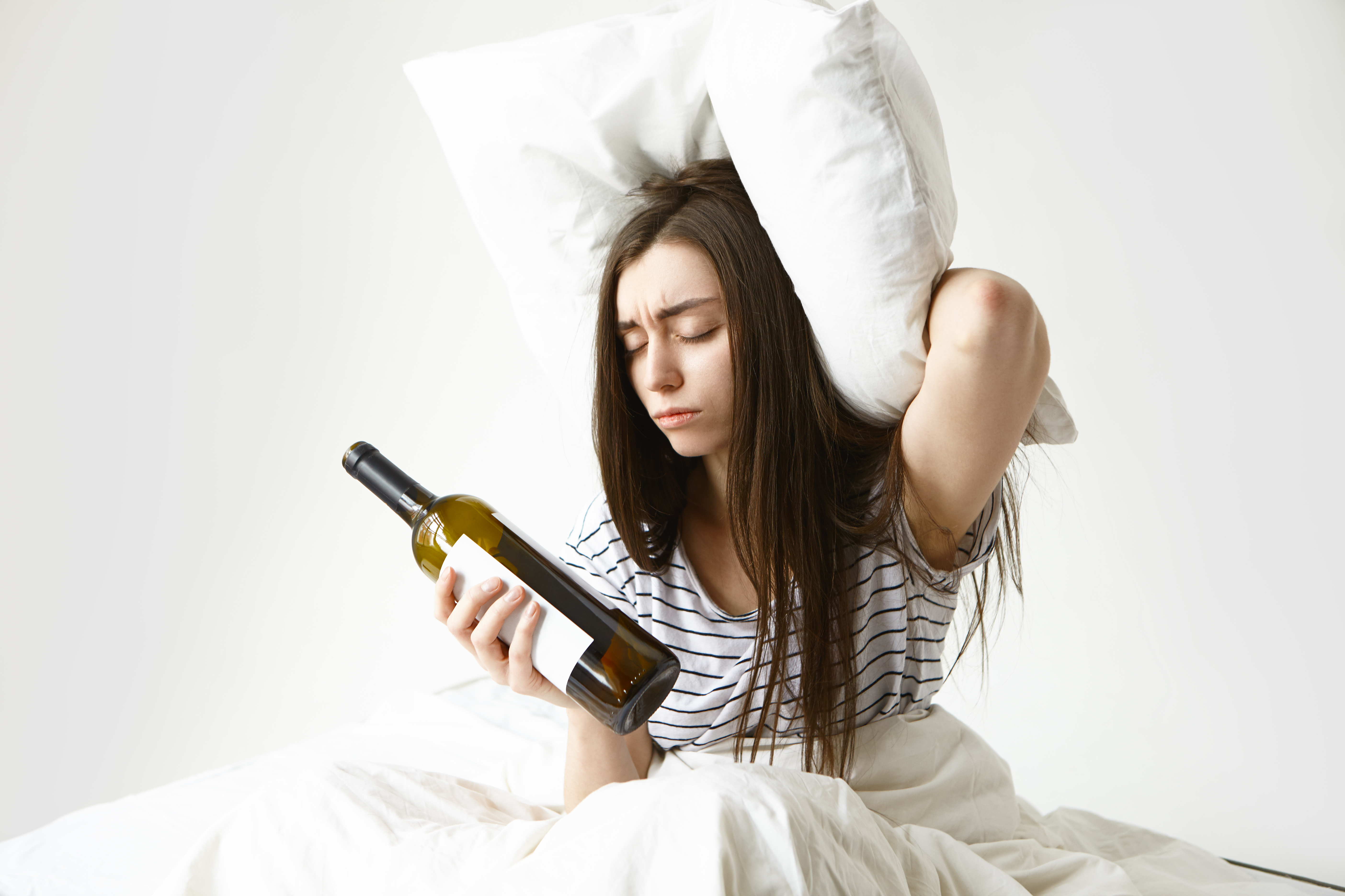 stressed-unhappy-woman-with-messy-loose-hair-suffering-from-headache-after-party-holding-pillow-empty-bottle