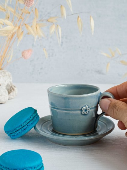 Modrý keramický šálek s podšálkem na dvojité espresso s reliéfem pomněnky držený v ruce vedle tyrkysových makronek