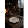 A rustic wooden table set in a cozy log cabin dining room, bathed in the warm glow of a crackling fireplace A white, snowflake patterned plate with the inscription 'Vánoční kalorie se nepočítají' sits promine