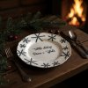 Close up lifestyle shot of a rustic wooden table set for a festive Christmas Eve dinner in a cozy, warmly lit cabin A white ceramic plate with delicate black snowflake designs and Czech script sits prominentl