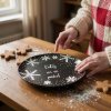 A close up lifestyle shot of a beautifully decorated, black ceramic plate with white snowflakes and elegant lettering on a rustic wooden table during a winter holiday baking session The scene features soft, d