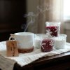 Close up lifestyle shot, a cozy winter scene featuring a steaming mug with a snowflake pattern, paired with handcrafted candles in glass jars, also adorned with snowflake designs, resting on a rustic wooden t