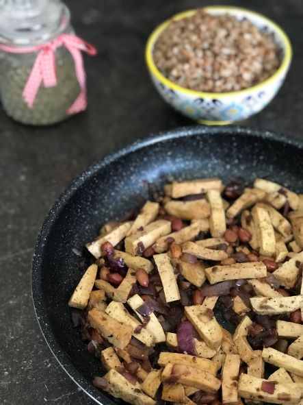 tofu na arašídech v pánvi, v pozadí vařená pohanka a skleněná nádobka s pepřem