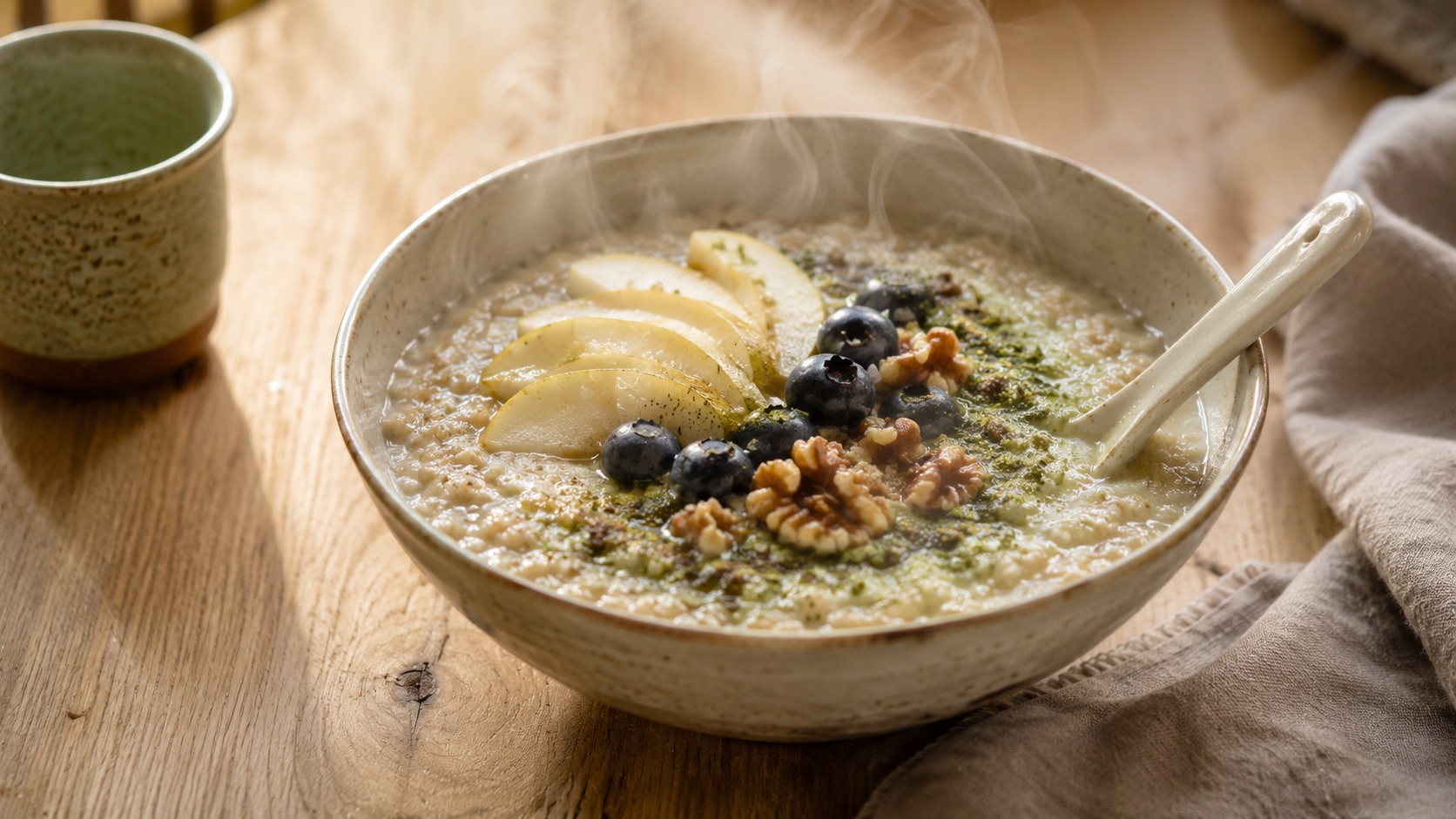Miska teplej pohánkovej kaše na raňajky ozdobená plátkami hrušky, čučoriedkami a orechmi. Nad kašou stúpa jemná para. Do kaše je zamiešaná štipka zeleného matcha prášku.