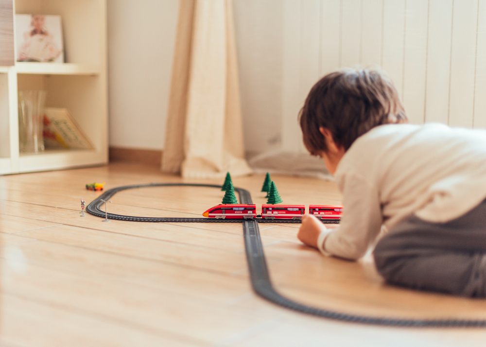 Dítě si hraje s dřevěnou vláčkodráhou na podlaze a sleduje elektrický vláček jedoucí po kolejích. Vláčkodráha s mostem, tunely a doplňky je ideální hračka pro rozvoj kreativity a motoriky.