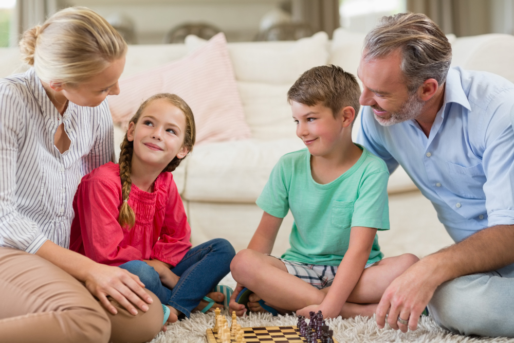 Rodiče a dvě děti sedí doma na koberci a společně hrají šachy