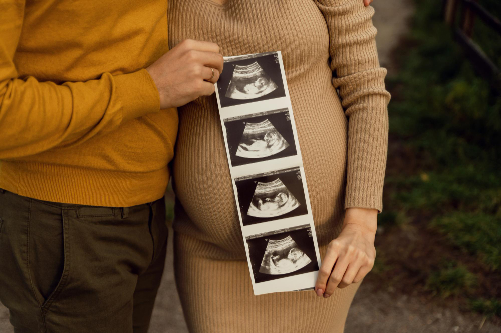 Těhotná žena se svým partnerem drží v ruce sérii snímků z ultrazvuku – symbol oznámení těhotenství rodičům a celé rodině.