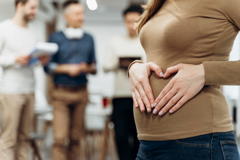 Těhotná žena v kanceláři ukazuje na bříško ve tvaru srdce, zatímco kolegové v pozadí diskutují – symbol oznámení těhotenství v práci.