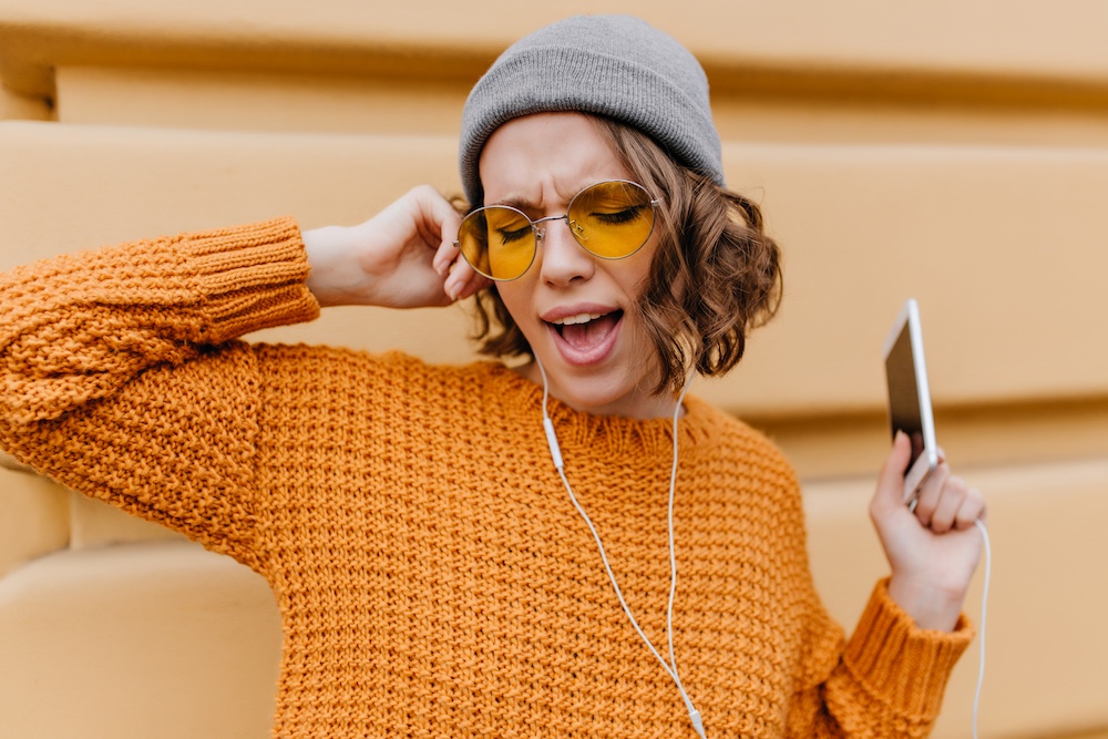funny-female-model-with-short-curly-hair-singing-favorite-song-while-standing-street-with-smartphone