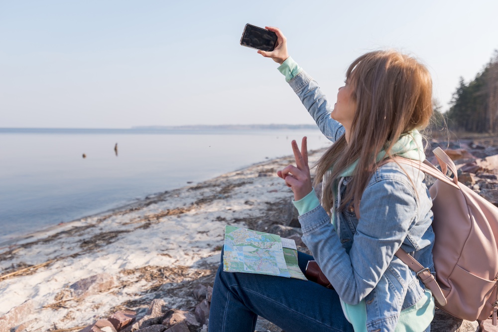 female-traveler-sitting-beach-taking-selfie-mobile-phone
