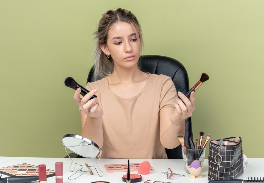 thinking-young-beautiful-girl-sitting-desk-with-makeup-tools-holding-looking-makeup-brush-isolated-olive-green-background_141793-119356