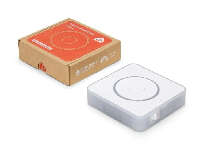 Home Assistant Voice Preview Edition vedle originální kartonové krabice, bílé čtvercové zařízení se zaoblenými rohy, průsvitnými boky a velkým kruhovým ovládacím prvkem na horní straně