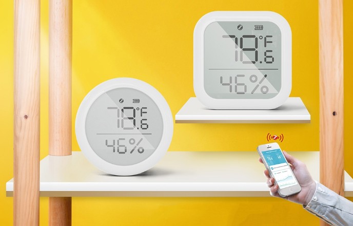Senzor teploty a vlhkosti MOES ZSS-KB-TH-LF-C Zigbee ve dvou provedeních – kulatý a čtvercový senzor s LCD displejem; vedle nich je mobilní telefon s aplikací pro sledování naměřených hodnot