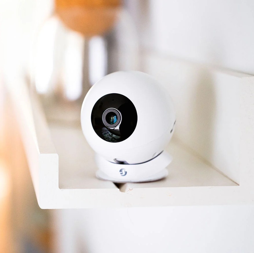 Bílá kamera Shelly Camera White umístěná na polici v interiéru s objektivem namířeným do místnosti