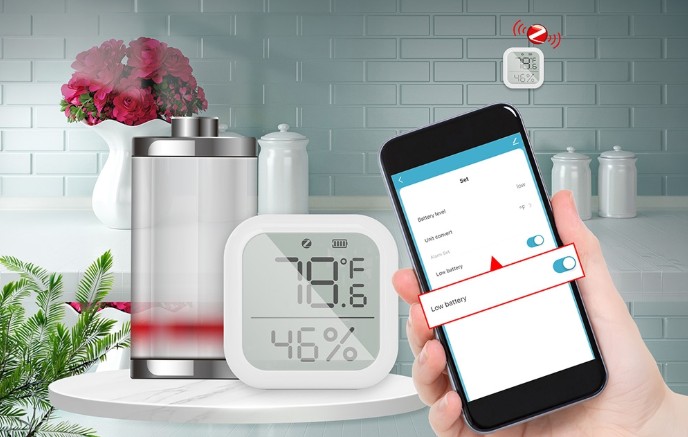 Senzor teploty a vlhkosti MOES ZSS-KB-TH-LF-C Zigbee umístěný v kuchyni vedle nádoby s květinami; LCD displej zobrazuje teplotu 19,6 °C a vlhkost 46 %, zatímco mobilní telefon zobrazuje upozornění na nízkou úroveň baterie