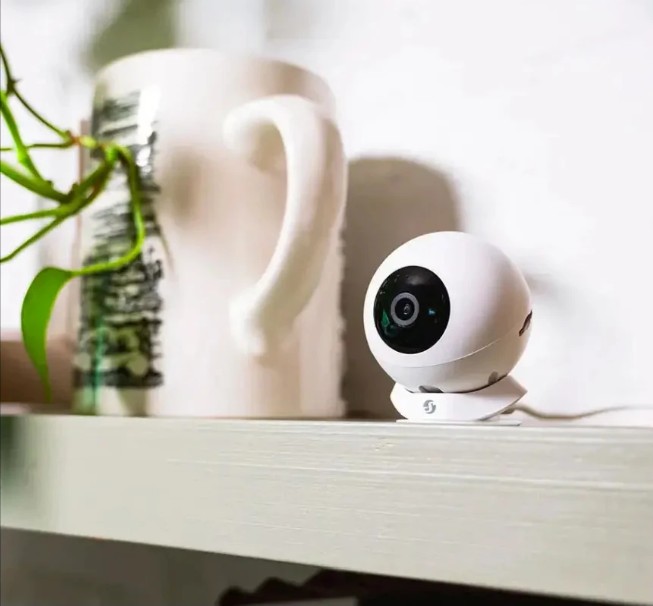 Bílá vnitřní kamera Shelly Camera White umístěná na bílé polici vedle hrnku a pokojové rostliny, s objektivem namířeným do prostoru