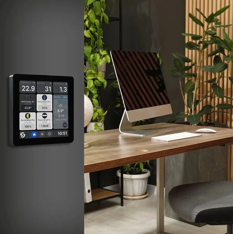 Shelly Wall Display X1i černý umístěný na stěně vedle pracovního stolu s počítačem. Displej slouží jako centrální ovládací panel chytré domácnosti a zobrazuje aktuální hodnoty a stav připojených zařízení.