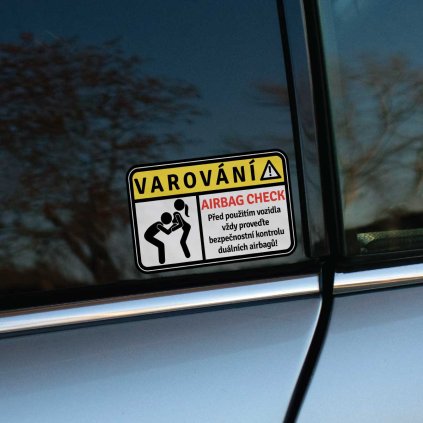 Tlačená nálepka v štýle výstražného štítku s textom „VAROVÁNÍ – AIRBAG CHECK“ a piktogramom dvoch postáv v humornom kontexte.