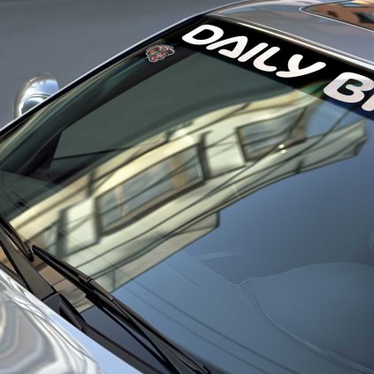 Sunstrip s veľkým bielym nápisom DAILY BITCH v stredovej časti v detaile na čelnom skle strieborného BMW.