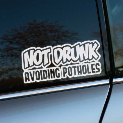 Vtipná biela nálepka s textom „Not Drunk, Avoiding Potholes“ je nalepená na okne auta. Tento dizajn humorne vysvetľuje, že neobvyklé manévrovanie auta je spôsobené snahou vyhnúť sa výtlkom, a nie konzumáciou alkoholu. Výrazné písmo s hrubým obrysom robí nálepku dobre viditeľnou a atraktívnou. Ideálna pre vodičov, ktorí chcú pridať trochu humoru na svoje vozidlo a pobaviť ostatných účastníkov cestnej premávky.