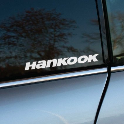 Biela nálepka s logom "Hankook" na zadnom okne automobilu s tmavými sklami. V pozadí sa odráža jemný obraz jesenných stromov, čo vytvára zaujímavý kontrast medzi čistým dizajnom nálepky a okolitou prírodou. Nálepka je ideálna pre milovníkov áut a fanúšikov značky Hankook, ktorí chcú zvýrazniť svoj štýl a podporu obľúbenej značke pneumatík. Výborne ladí s metalickou sivou karosériou auta a pridáva vozidlu športový charakter.  **Kľúčové slová:** nálepka na auto Hankook, nálepky na okno auta, tuningové nálepky, reklamná nálepka na auto, športové auto doplnky, dizajnové nálepky, polepy na auto, metalická sivá karoséria, kvalitné auto nálepky, nálepky značky pneumatík.