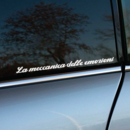 Plotrovaná nálepka s nápisom „La meccanica delle emozioni“ v elegantnom skriptovom štýle, slogan značky Alfa Romeo.