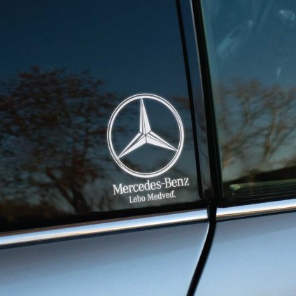 Fotografia zobrazuje detail okna auta Mercedes-Benz s nalepenou nálepkou, ktorá obsahuje logo značky a slogan "Lebo Medveď." Táto vtipná variácia na klasický slogan pridáva autu osobitý a originálny nádych, ideálny pre majiteľov, ktorí chcú prejaviť svoju hrdosť na značku Mercedes-Benz so štipkou humoru. Nálepka elegantne ladí s tmavým pozadím, čo z nej robí štýlový doplnok pre každé vozidlo tejto značky.