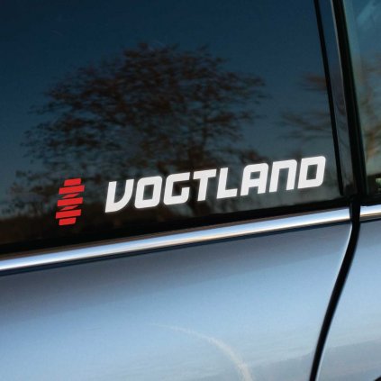 Biela nálepka "VOGTLAND" s moderným písmom a výrazným červeným symbolom pružiny na zadnom bočnom okne strieborno-sivého auta. Táto nálepka zdôrazňuje spojenie so špičkovými podvozkovými systémami a športovým tuningom, pričom vyniká na tmavom tónovanom skle. Ideálna pre automobilových nadšencov, ktorí chcú vyjadriť svoj štýl a podporu kvalitného vybavenia.  **Kľúčové slová:** nálepka na auto, samolepka VOGTLAND, tuningová nálepka, logo VOGTLAND, nálepka na sklo auta, športové nálepky, automobilové nálepky, personalizácia auta, doplnky na auto, podvozkové systémy, tuning auta.