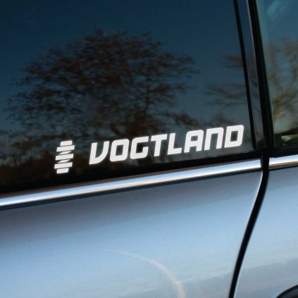 Biela nálepka "VOGTLAND" s moderným písmom a symbolom pružiny na zadnom bočnom okne strieborno-sivého auta. Dizajn nálepky zdôrazňuje spojenie so špičkovým podvozkom a športovým tuningom, pričom vyniká na tmavom tónovanom skle. Perfektný doplnok pre fanúšikov automobilového tuningu a kvalitných odpružení.  **Kľúčové slová:** nálepka na auto, samolepka VOGTLAND, tuningová nálepka, logo VOGTLAND, nálepka na sklo auta, automobilové nálepky, personalizácia auta, športové nálepky, doplnky na auto, podvozkové systémy.