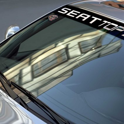 Detailný pohľad na čelné sklo strieborného BMW s nápisom "SEAT TOP SPORT" a malou nálepkou maskota. Odrážajúce sa budovy na skle pridávajú hĺbku a mestský charakter.