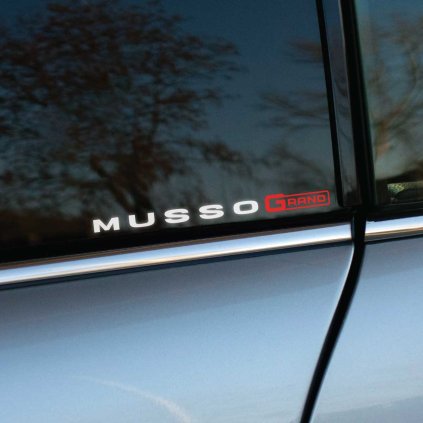 Plotrovaná nálepka s nápisom „MUSSO GRAND“, kde slovo „GRAND“ je zvýraznené červenou farbou. Ideálny doplnok pre majiteľov SsangYong Musso Grand.